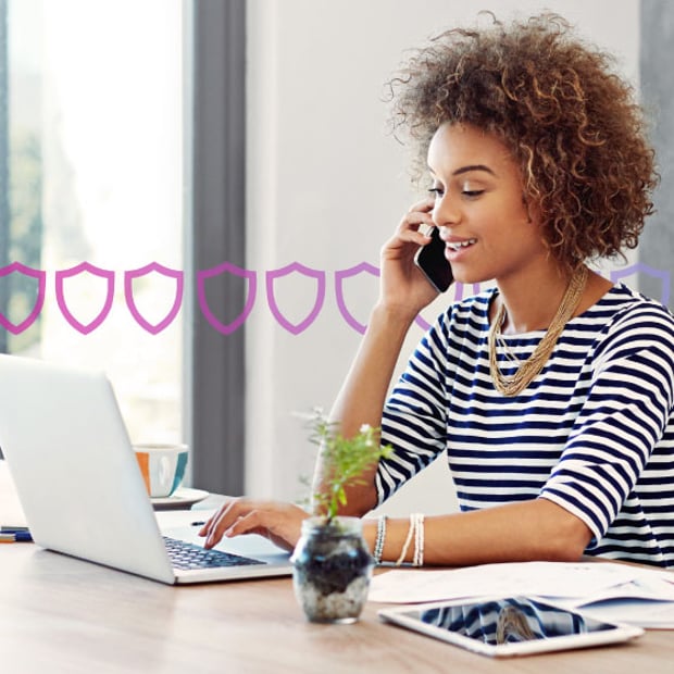 UC UCaaS Security Blog Header Photo of a woman in an office setting talking on her phone while typing or scrolling on her laptop. In the background, a series of small purple shields runs across the frame, representing security.