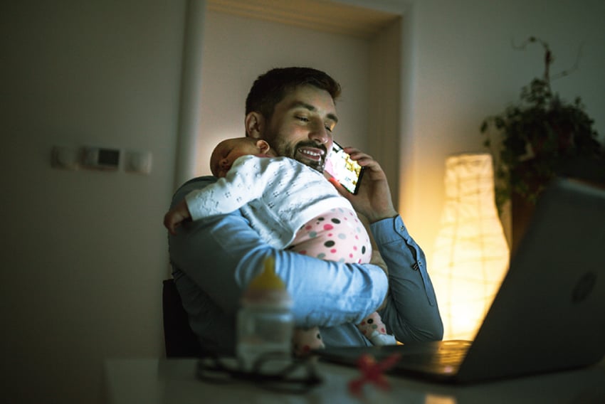 Photo of a business man at home, talking on his cellphone. He is holding a sleeping infant over his shoulder.