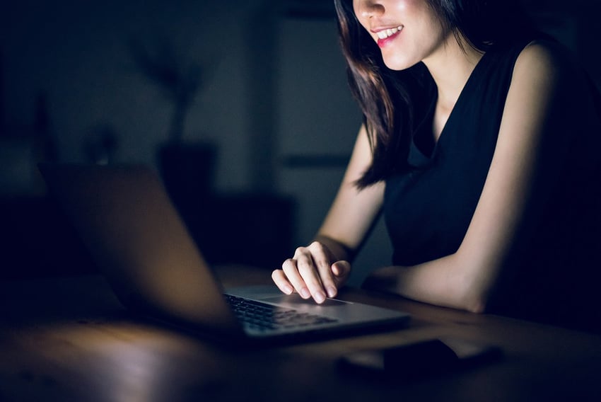 Image of woman smiling and sitting in a dark room on her laptop 