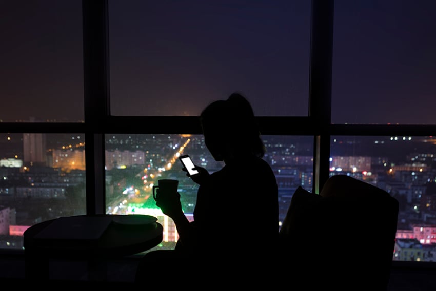 Photo of a woman in an office at night. The lights are out, and we can see her in the glow of her cellphone and the lights of a city outside her window.
