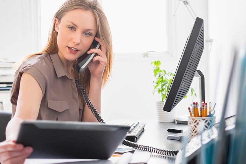 Woman sitting at a desk, on the phone, looking at a tablet. This conveys VoIP phone service.