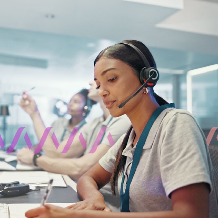 Photo of several contact center employees sitting side by side. The woman in the foreground is wearing a headset and writing something on a notepad.
