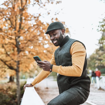 Photo of a smiling man standing outdoors. He is looking at something on his phone.