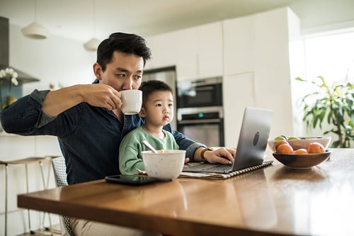 man and son are in a kitchen looking at a laptop together, father is holding a coffee cup