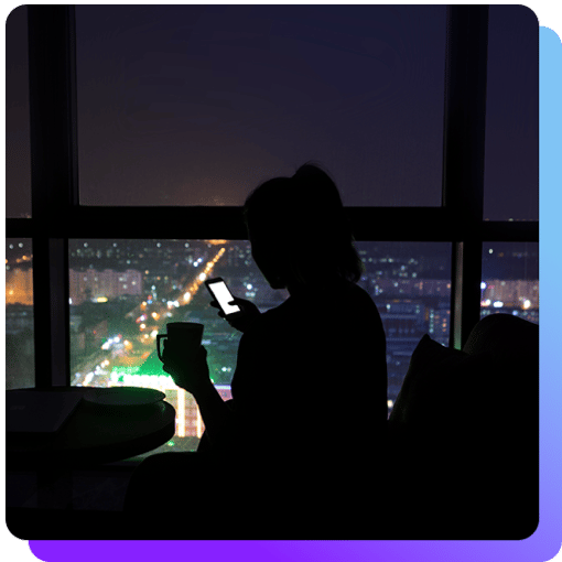 Silhouette of a woman looking at her phone with coffee in hand with a night time cityscape below