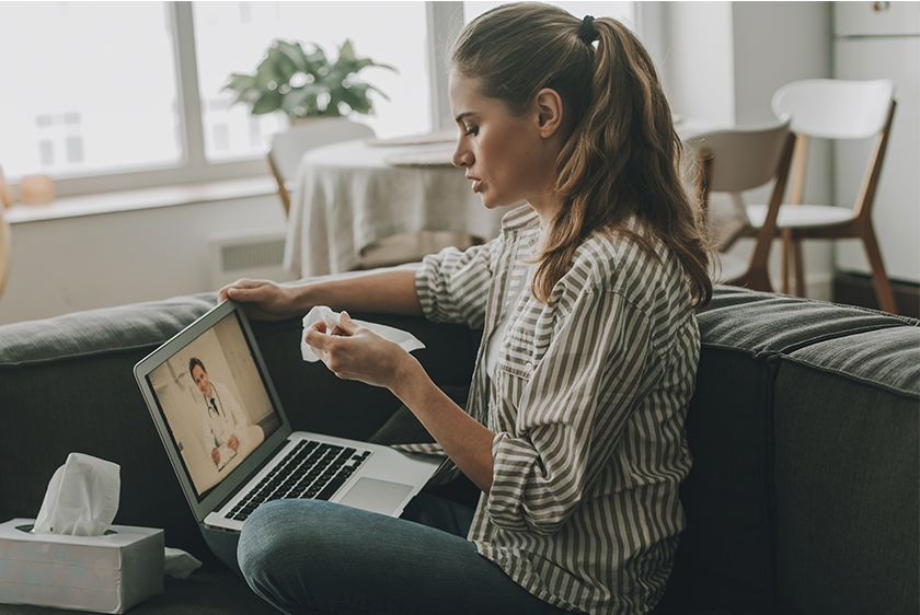 Photo of a woman sitting on a couch with her laptop open. On the screen we can see a doctor who the woman is having a consultation with.
