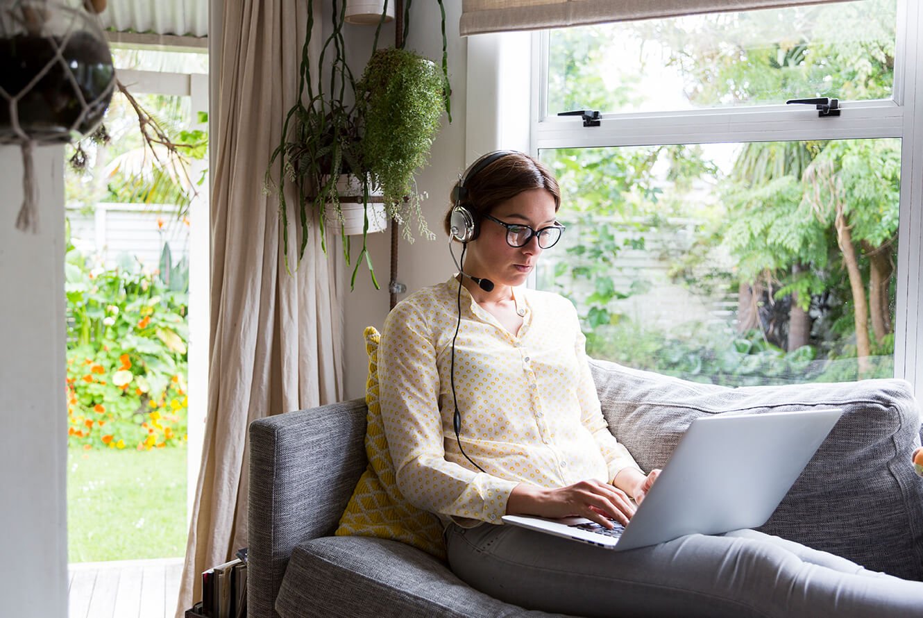 Woman sitting on her coach at home working from her laptop with headset on